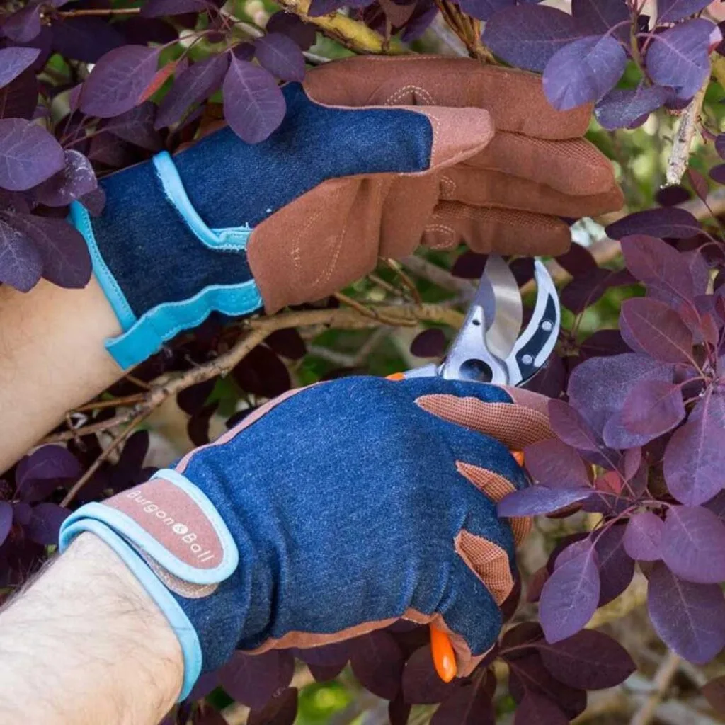 A gardener wearing denim gardening gloves with leather trim while pruning a tree with secateurs.