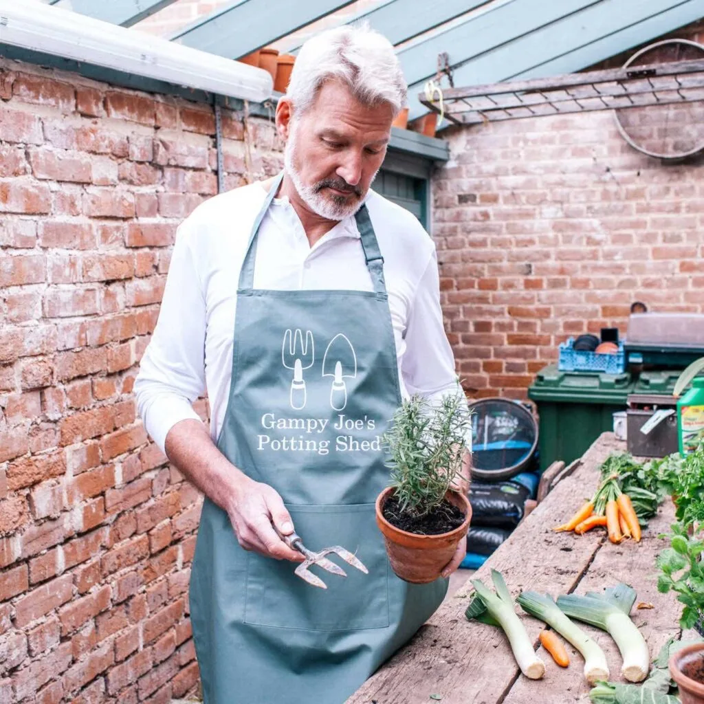 A man wearing a grey apron holding pot of rosemary in a potting shed.